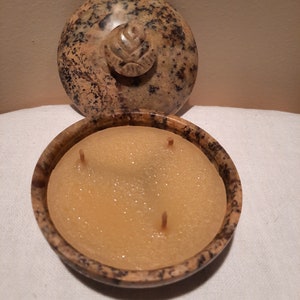 May include: A round, speckled stone candle holder with a lid. The lid features a carved floral design. The candle inside is a light yellow color with three wicks. The candle holder is open, revealing the candle.