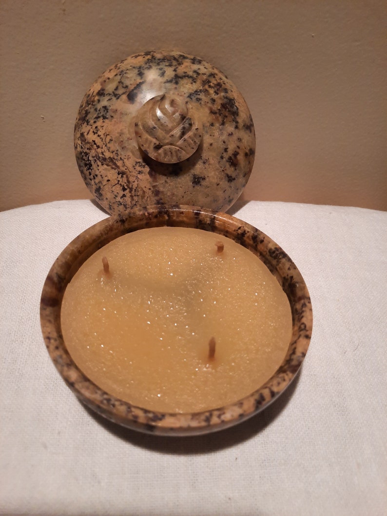 May include: A round, speckled stone candle holder with a lid. The lid features a carved floral design. The candle inside is a light yellow color with three wicks. The candle holder is open, revealing the candle.