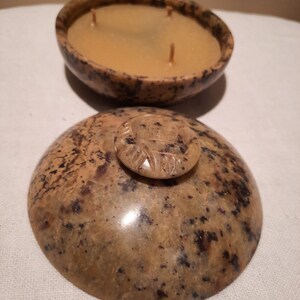 May include: A natural stone candle with a speckled brown and tan pattern. The candle has three wicks and a light brown wax. The lid, also stone, features a small, carved handle. The candle is in a bowl-shaped container.