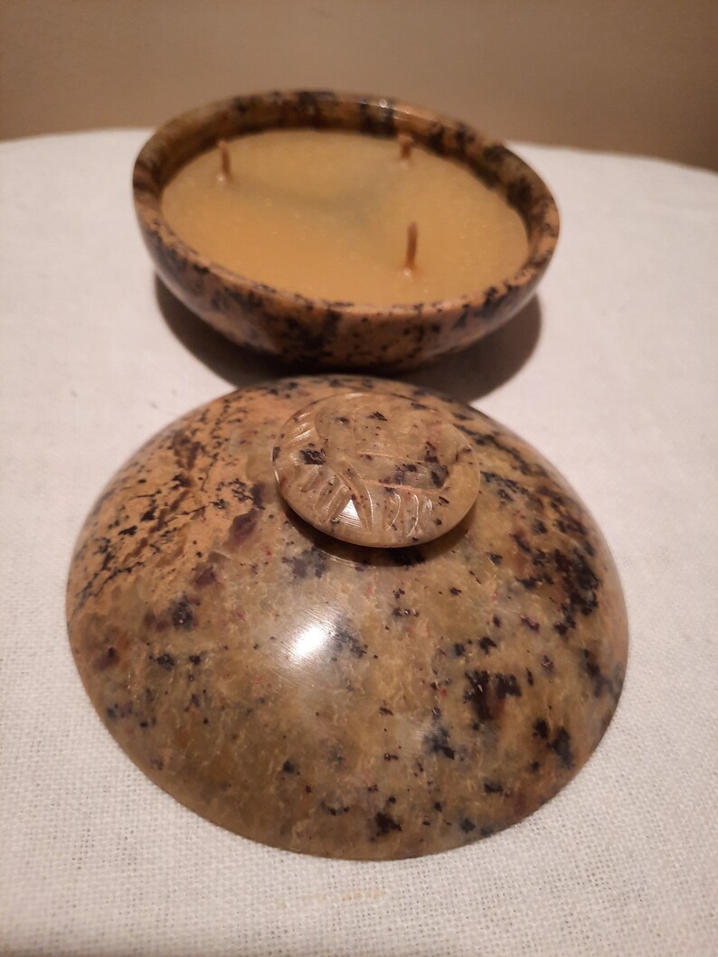 May include: A natural stone candle with a speckled brown and tan pattern. The candle has three wicks and a light brown wax. The lid, also stone, features a small, carved handle. The candle is in a bowl-shaped container.