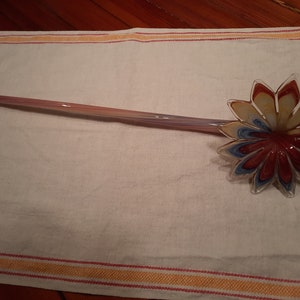 May include: A decorative glass stirring spoon with a flower-shaped head. The petals are red, yellow, and blue. The handle is a light pink and purple colour. The spoon is resting on a white towel.