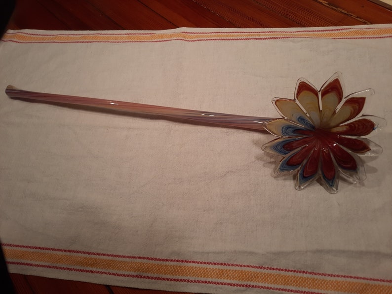 May include: A decorative glass stirring spoon with a flower-shaped head. The petals are red, yellow, and blue. The handle is a light pink and purple colour. The spoon is resting on a white towel.
