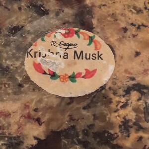 May include: A close-up of a round, slightly damaged sticker with the words "Krishna Musk" in black text. The sticker is decorated with red and orange floral accents and the name "R. Esopo" in small black text.