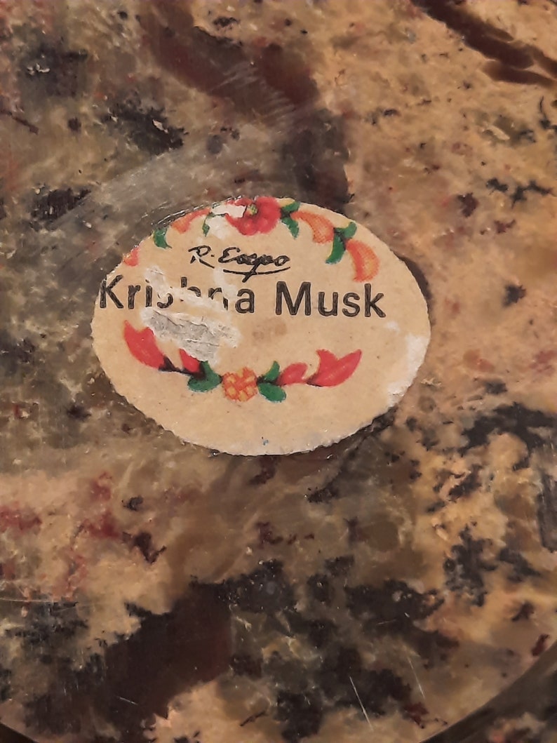 May include: A close-up of a round, slightly damaged sticker with the words "Krishna Musk" in black text. The sticker is decorated with red and orange floral accents and the name "R. Esopo" in small black text.