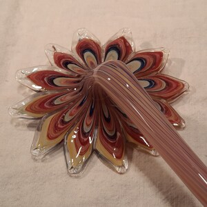 May include: A handmade glass flower with a long stem. The flower petals are a mix of red, yellow, and blue, with a clear glass edge. The stem is a light purple colour with vertical stripes.