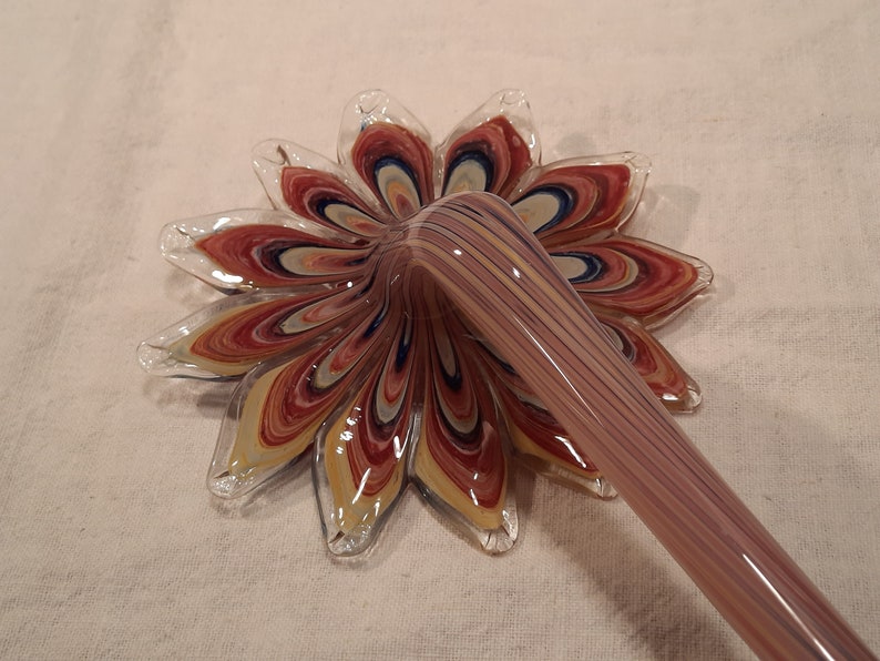 May include: A handmade glass flower with a long stem. The flower petals are a mix of red, yellow, and blue, with a clear glass edge. The stem is a light purple colour with vertical stripes.