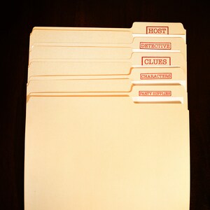 May include: A collection of manila file folders with red text labels. The labels read "HOST", "DETECTIVE", "CLUES", "CHARACTERS", and "PARTY SUPPLIES". The folders are placed on a dark brown surface.