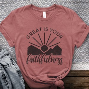 Może przedstawiać: T-shirt z krótkim rękawem w kolorze przybrudzonego różu z ciemnobrązową grafiką przedstawiającą słońce wschodzące nad szczytami gór. Napis "GREAT IS YOUR" jest wydrukowany pogrubioną czcionką bezszeryfową nad słońcem, a "faithfulness" kursywą.