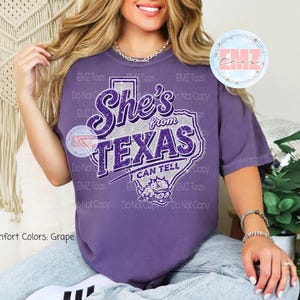 May include: A grape-coloured t-shirt featuring a graphic of the state of Texas in silver glitter. The text reads She's from TEXAS I CAN TELL in purple glitter script and block lettering. A small purple graphic of a horned lizard is below the text.