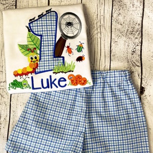 以下が含まれることがあります： 白地に青と白のチェック柄の数字1、虫眼鏡、毛虫、コオロギ、クモが刺繍された白いTシャツ。シャツには「Luke」という名前も刺繍されています。シャツは青と白のチェック柄のショートパンツと組み合わせています。