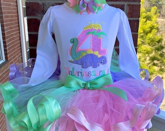 Könnte beinhalten: Ein weißes Langarmshirt mit einem rosa-grünen Dinosaurier-Applikationen und dem Text "Valeriasaurus 4". Das Shirt wird mit einem rosa-grünen Tüll-Tutu mit passenden Bändern kombiniert.