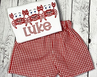 Könnte beinhalten: Ein weißes T-Shirt mit roten gestickten Krabben und dem Namen "Luke" auf der Vorderseite. Das Shirt wird mit rot-weiß karierten Shorts kombiniert.