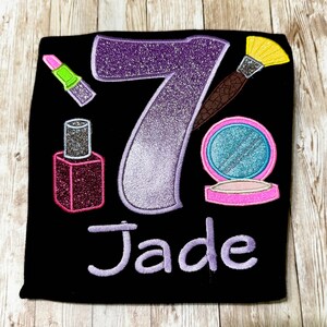 Pode incluir: Uma camiseta preta com um número 7 roxo brilhante, um estojo de maquiagem rosa e prateado, um pincel de maquiagem marrom com cabeça de pincel amarela, um frasco de esmalte de unha rosa e prateado, um batom verde e rosa, e o nome "Jade" em purpurina roxo.