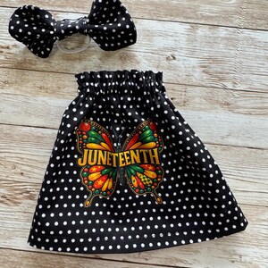Può includere: Una gonna a pois bianchi e neri e un fiocco abbinato. La gonna presenta un colorato disegno a farfalla con la scritta "Juneteenth" in lettere dorate. Il fiocco è attaccato a un elastico trasparente.