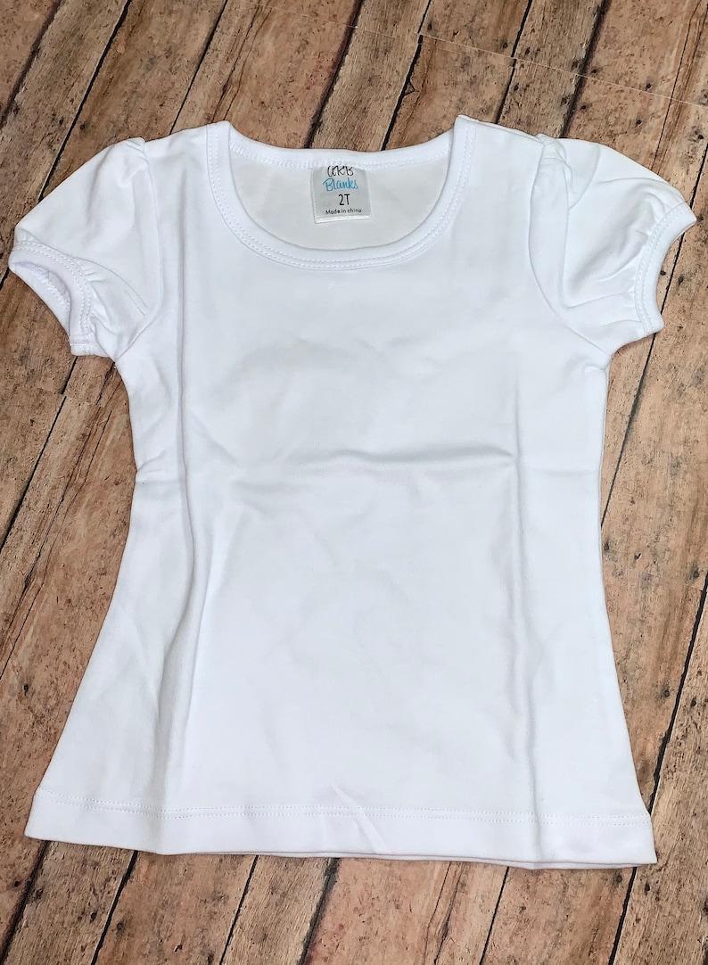 Peut inclure: T-shirt blanche &agrave; manches courtes avec un col rond. Le t-shirt a une petite &eacute;tiquette sur le devant qui dit "Little Blanks 2T Made in China".