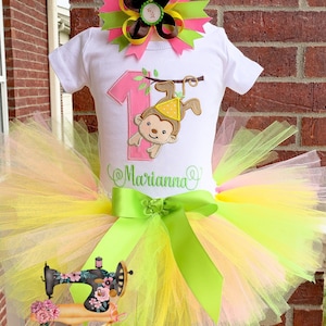 Puede incluir: Un tutú de tul rosa, amarillo y verde con un lazo de satén verde. Un body blanco con un número uno rosa y un mono colgado de una rama. El body está personalizado con el nombre "Marianna". Un lazo rosa, amarillo y verde con un centro marrón está en la parte superior del body.