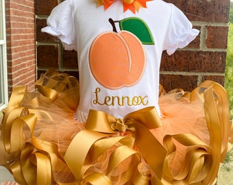 Peut inclure: Un t-shirt blanc avec un motif de pêche brodé et le nom "Lennox" en dessous. Le t-shirt est associé à un tutu en tulle avec des accents de ruban de satin doré et un nœud pêche et or.