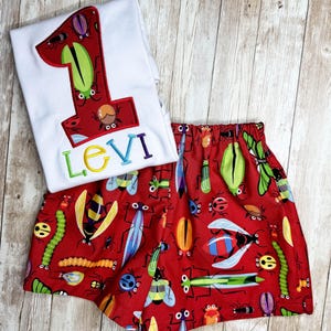 以下が含まれることがあります： 様々な昆虫のデザインで飾られた赤い数字の「1」のアップリケが付いた白い半袖シャツと、カラフルな文字で刺繍された名前「LEVI」。カラフルな昆虫模様の赤いショートパンツと組み合わせています。