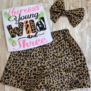May include: A white t-shirt with pink and brown embroidered text reading Heiress Young wild and Three, accented with green palm leaves. The set includes leopard print shorts and a matching leopard print hair bow with a central knot detail.
