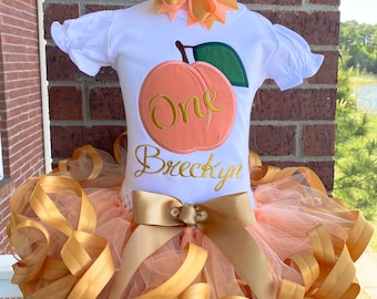 Peut inclure: Tenue de premier anniversaire sur le thème de la pêche pour une fille. Un t-shirt blanc avec un motif de pêche et le mot "One" brodé en or. Le t-shirt porte également le nom "Breckyn" brodé en or. La tenue comprend un tutu en tulle pêche et or avec un nœud en satin doré.