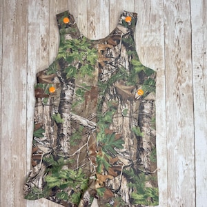 May include: Camouflage print baby overalls with orange buttons. The overalls are made of a soft, comfortable fabric and have a relaxed fit.