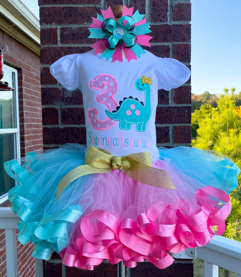 Peut inclure: Un t-shirt blanc avec un chiffre 3 rose et un dinosaure turquoise &agrave; pois roses. Le t-shirt dit "Shannahsarus" et "3" avec un n&oelig;ud jaune sur le dinosaure. Le t-shirt est associ&eacute; &agrave; un tutu en tulle turquoise et rose avec un ruban dor&eacute;.