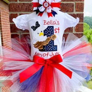 Pode incluir: Um tutu branco com camadas de tule vermelho, branco e azul. Um laço de cetim vermelho é amarrado na cintura. Uma camiseta branca com um laço vermelho, branco e azul, uma águia e o texto "Bella's 1st 4th of July" é usado sobre o tutu.