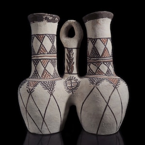 May include: A white ceramic triple-spouted vase with a geometric pattern in brown and black. The vase has a central handle and a unique shape.
