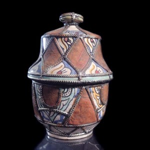 May include: A decorative ceramic jar with a lid. The jar features a brown leather-like texture with diamond-shaped metal accents. The lid and body are adorned with colorful floral and geometric patterns, including blue, yellow, and white designs. The jar has a silver-colored metal trim.