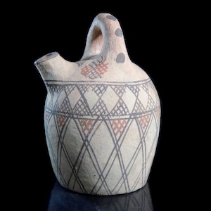 May include: A cream-colored ceramic pitcher with a handle and spout. The pitcher is decorated with geometric patterns in black, red, and gray. The design includes diamond and crosshatch motifs. Two gray dots are on the top of the handle.