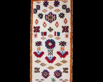 Può includere: Un tappeto rettangolare bianco sporco con un vivace motivo geometrico. Il design presenta motivi ripetuti in rosso, blu, verde e marrone, con un bordo arancione e bordi sfrangiati. I dettagli intricati e la tavolozza dei colori del tappeto creano un tessuto visivamente accattivante.