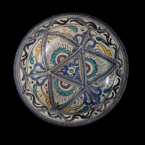 Op de afbeelding: Een ronde keramische kom met een handgeschilderd ontwerp met een centraal stermotief. Het oppervlak toont ingewikkelde patronen in tinten blauw, groenblauw, geel en donkerbruin op een crèmekleurige achtergrond, met een textuurrijke, donkere rand.