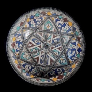 Puede incluir: Un plato decorativo redondo con un diseño geométrico detallado. El plato presenta un patrón de estrella central con intrincados azulejos en azul, amarillo, marrón y turquesa. Un borde de metal plateado rodea el plato.