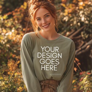 May include: A long-sleeved olive green shirt with the text "YOUR DESIGN GOES HERE" in white. The shirt is worn by a person with red hair and a bun. The background is blurred with autumn colors.