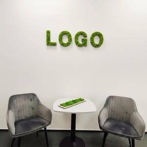 Puede incluir: Un espacio de oficina moderno con dos sillones de terciopelo gris y una pequeña mesa blanca. Una jardinera rectangular llena de musgo verde está sobre la mesa. La palabra "LOGO" está escrita con musgo verde en la pared blanca.