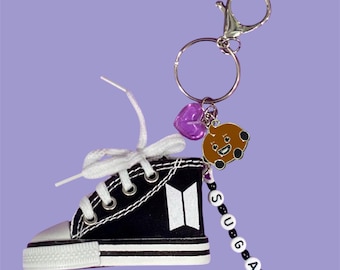May include: A black and white miniature sneaker keychain featuring a white BTS logo on the side. The keychain includes a silver metal ring, a translucent purple heart charm, a brown cartoon character charm, and a beaded strand spelling out SUGA.