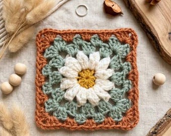 May include: A square crochet granny square featuring a central white daisy with a yellow center, surrounded by layers of sage green and terracotta orange yarn. The square is constructed with a classic cluster stitch pattern in a flat, square shape.