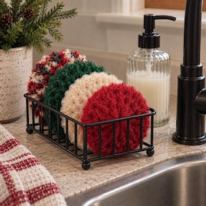 May include: A black metal rectangular wire rack holding four round, textured scrubbers. The scrubbers are arranged in a row and feature distinct colors: a red, white, and green variegated pattern, a solid dark green, a solid cream, and a solid red.