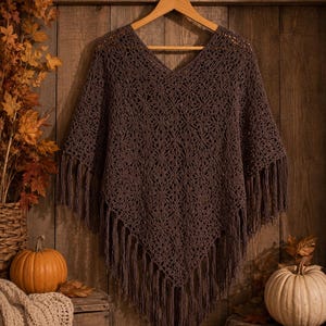 May include: A dark brown crochet poncho featuring an intricate star-like pattern throughout the fabric. The garment has a V-neckline and a pointed hemline finished with long, dangling fringe trim along the edges, displayed on a wooden hanger.