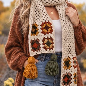 Puede incluir: Una bufanda larga de ganchillo con una base de color crema y motivos de cuadrados de la abuela multicolores en tonos amarillo mostaza, verde oliva, naranja quemado y marrón. Los extremos de la bufanda tienen borlas de lana gruesa a juego.
