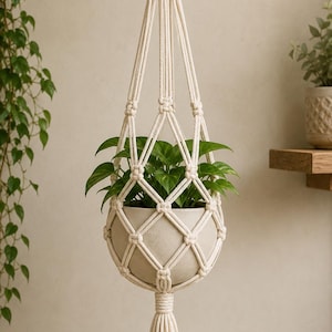 May include: A cream-coloured macrame plant hanger, featuring a woven design, suspending a round, light-coloured pot filled with green leafy plants. The hanger has a wooden ring at the top and a tassel at the bottom. The plant pot is approximately 15 cm in diameter.