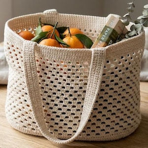 May include: A cream-coloured, crocheted tote bag filled with oranges and greenery. The bag has a rectangular shape with a wide opening and two handles. A book titled "The Paper Pal" is visible inside the bag. The bag is sitting on a wooden surface.