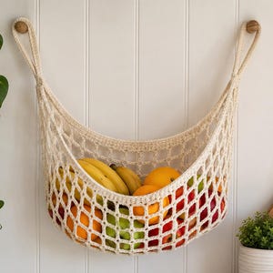 Puede incluir: Una hamaca de frutas de ganchillo color crema llena de plátanos, naranjas, manzanas y frutos rojos, colgada en una pared blanca. La hamaca está suspendida por perillas de madera. Una planta verde es visible en el lado izquierdo de la imagen.