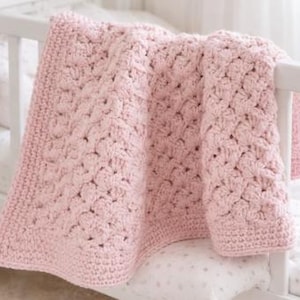 Puede incluir: Una manta de ganchillo rosa pálido sobre una barandilla blanca de cuna. La manta presenta un patrón de puntadas texturizadas y un borde liso. La manta probablemente está hecha de un hilo suave, para un bebé o niño.
