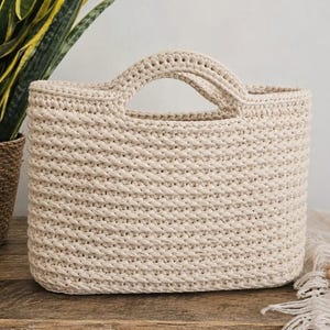 May include: A beige, rectangular crochet tote bag with two handles. The bag is made of thick yarn, with a textured stitch pattern. The bag is sitting on a wooden surface, with a plant and decorative beads in the background.