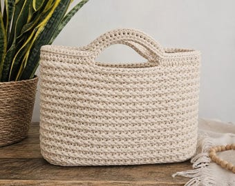May include: A beige, rectangular crochet tote bag with two handles. The bag is made of thick yarn, with a textured stitch pattern. The bag is sitting on a wooden surface, with a plant and decorative beads in the background.