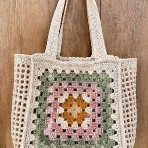 May include: Cream-colored crochet tote bag with a square, multi-colored floral design in the center. The floral design features shades of pink, green, and orange. The bag has two handles and a mesh-like pattern.