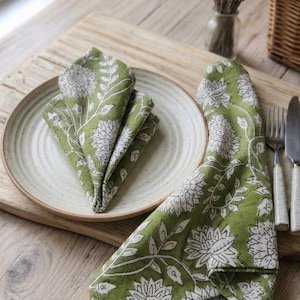 May include: Two green cloth napkins featuring a white floral and vine block print pattern. One napkin is folded into a triangle shape and placed on a round, off-white ceramic plate, while the other napkin is draped beside the plate.