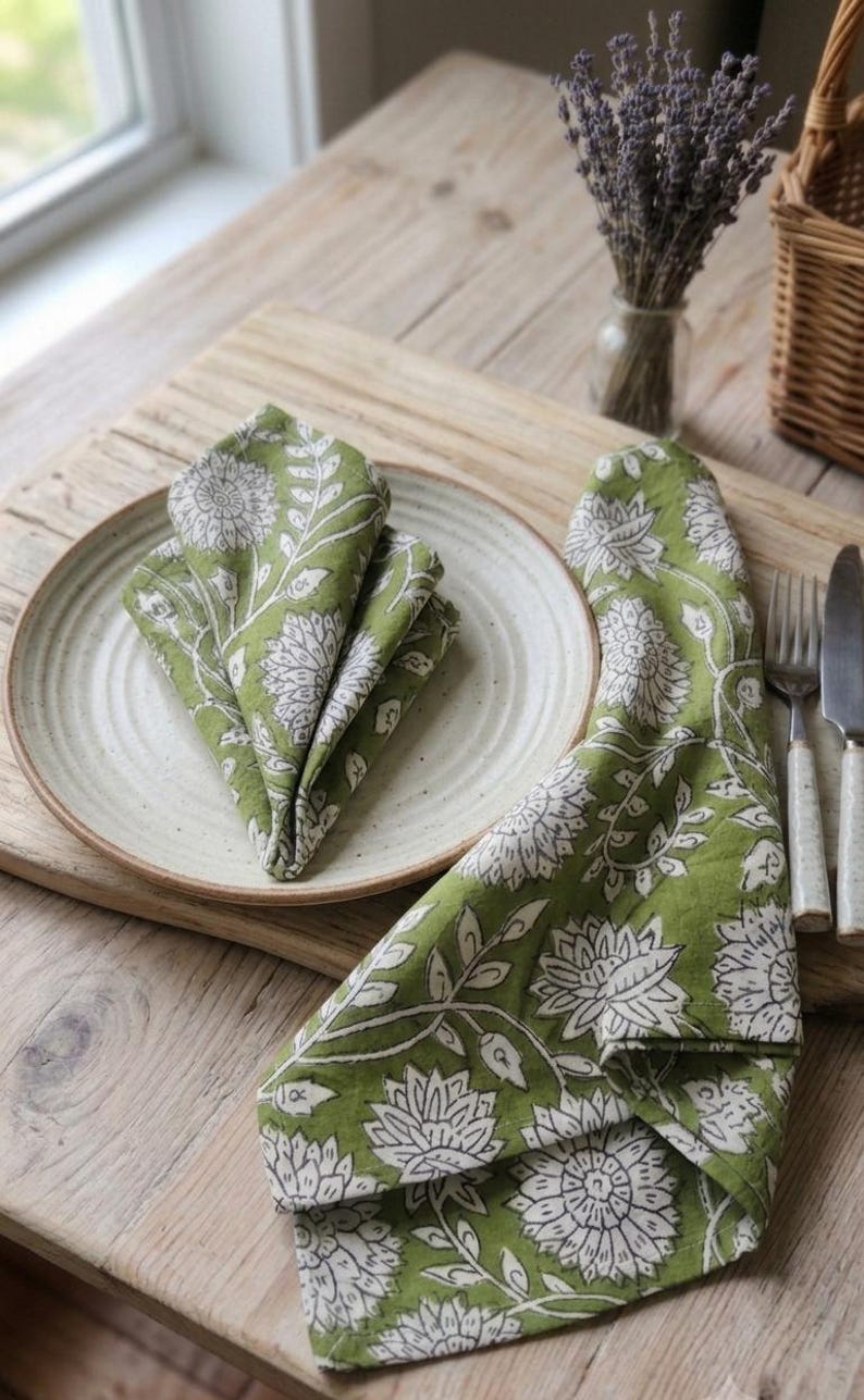 May include: Two green cloth napkins featuring a white floral and vine block print pattern. One napkin is folded into a triangle shape and placed on a round, off-white ceramic plate, while the other napkin is draped beside the plate.