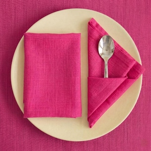 May include: A cream-coloured plate with a folded fuchsia napkin and a silver spoon. Another fuchsia napkin is placed to the left. The setting is on a fuchsia tablecloth.
